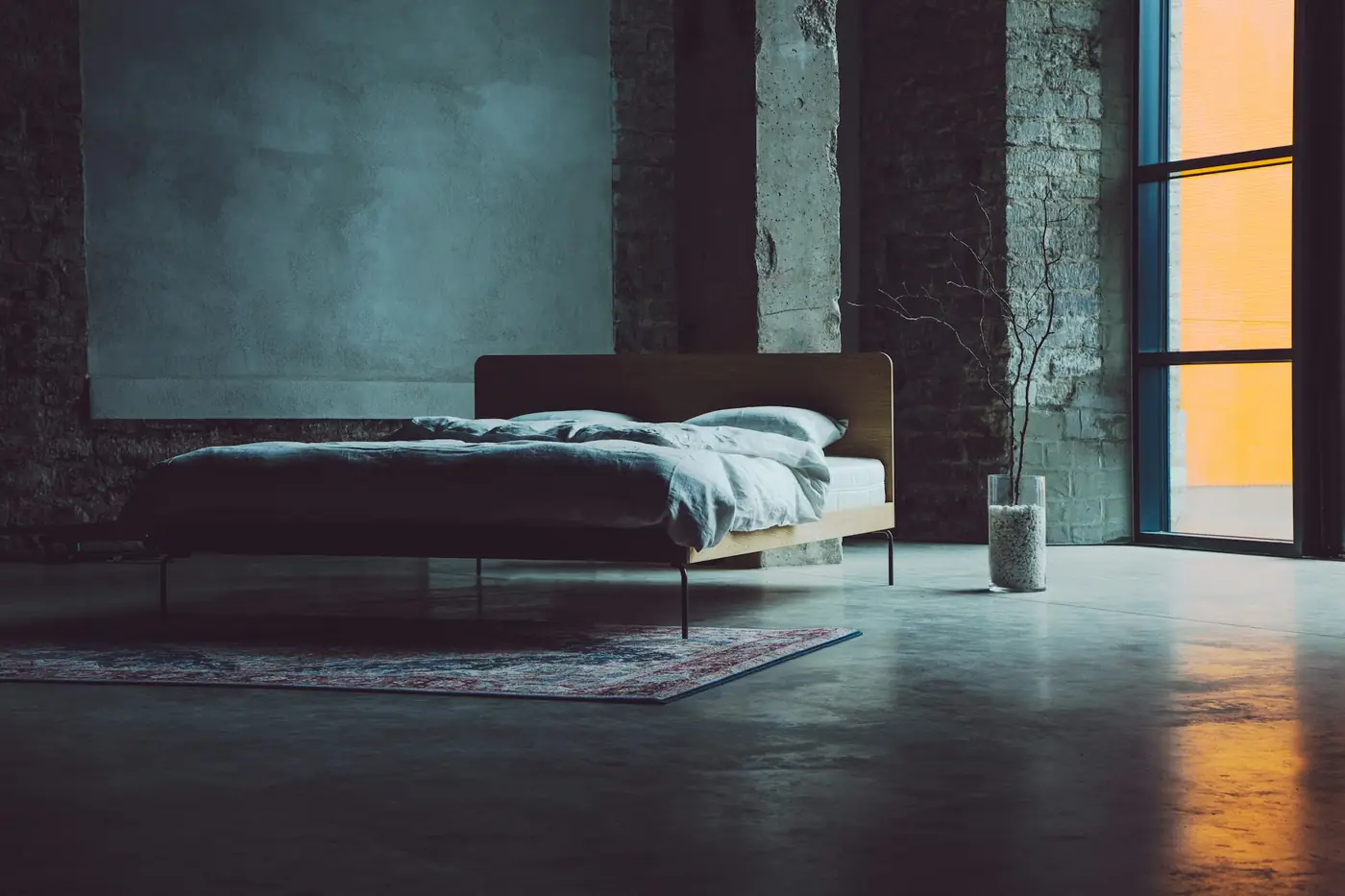 Quarto amplo e escuro, de paredes de concreto, com a cama arrumada e a luz quente do fim de tarde entrando pela janela